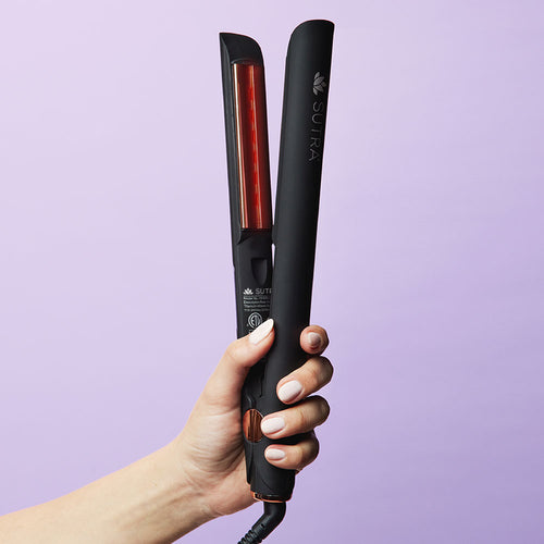 A black flat iron with gold color details and a infrared light inside, being held by a female hand on a light purple background